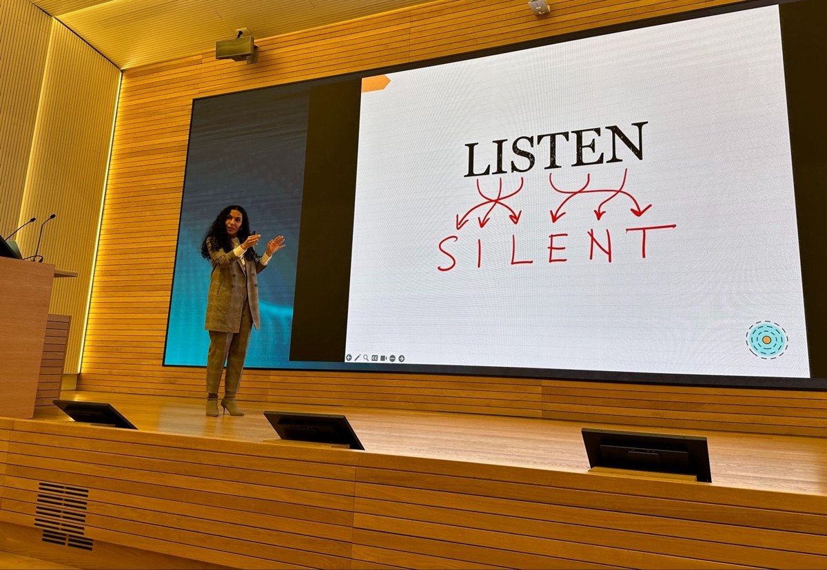 Aline speaking on stage beside a LISTEN and SILENT slide